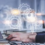 Hands of businessman typing on laptop in blurred office with double exposure of gears and graphs.